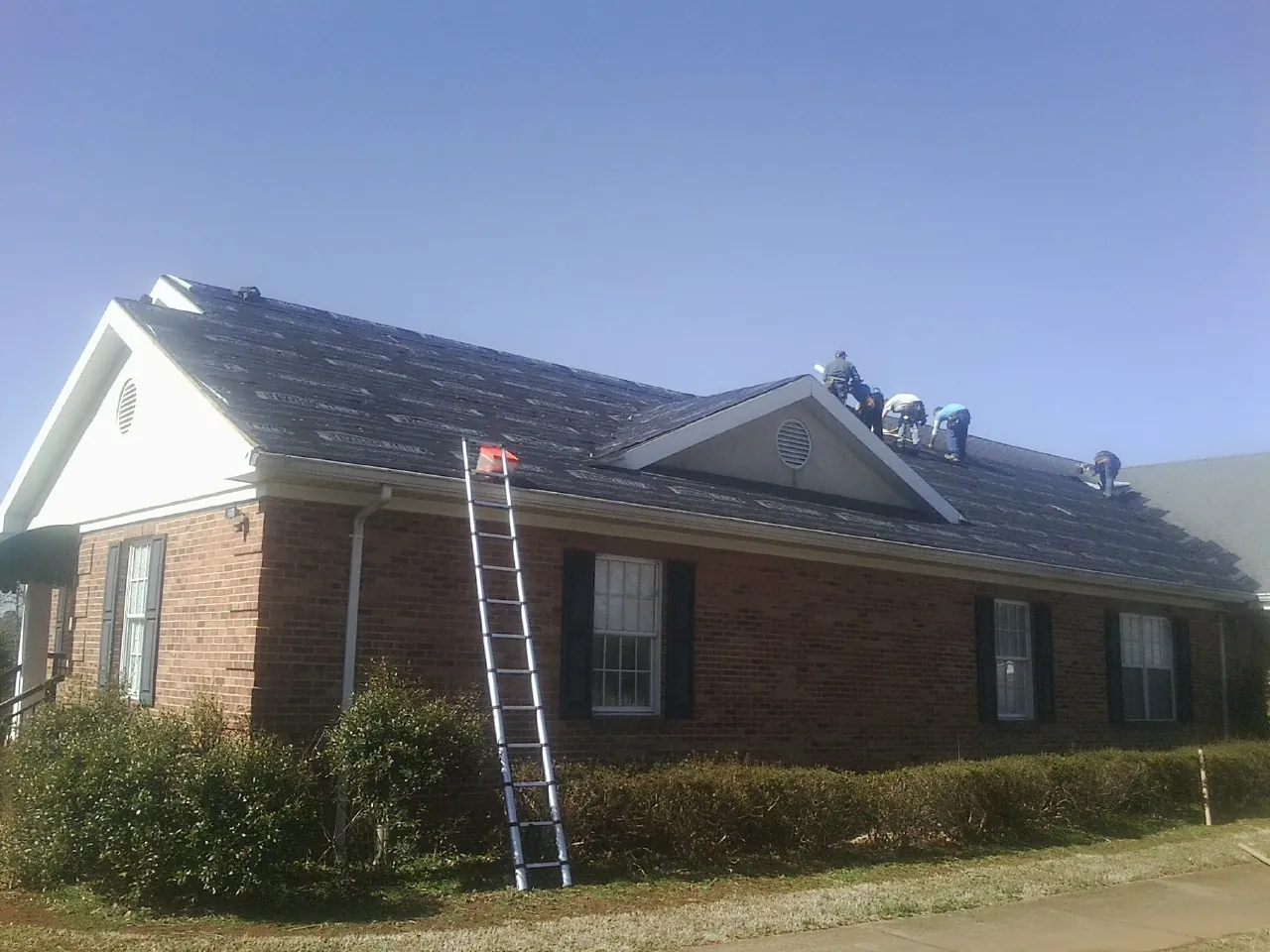 Crew tearing shingles off a brick ranch roof with a ladder set — Metal Roof Repair work by our crew in New England
