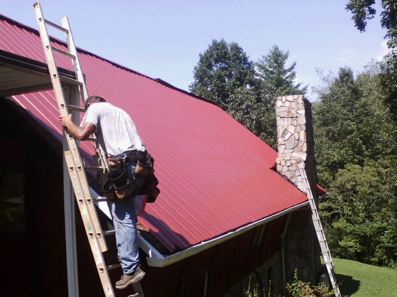 Roofer on a ladder beside a red metal roof and stone chimney — Soffit & Fascia Repair by Roofing Brothers in New England